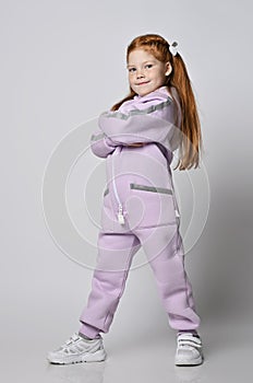 Cool self-confident kid girl in pink modern jumpsuit stands with arms crossed at chest, looking at camera