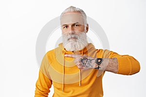 Cool senior hipster man with tattoos pointing left at copy space, showing advertisement, grandad with long beard wearing