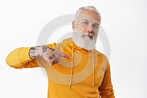 Cool senior man with tattoos pointing at himself. Bearded cool grandfather in orange hoodie self-promoting, bragging photo