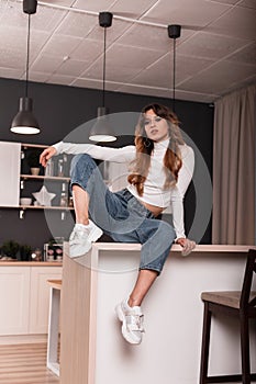 Cool sexy red-haired young woman in a stylish white top in fashionable blue jeans in trendy leather sneakers is resting on a