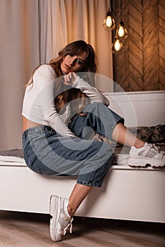 Cool sexy red-haired young woman in a stylish white top in fashionable blue jeans in trendy leather sneakers is resting on a soft