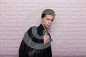 Cool sexy young man fashion model with a stylish hairstyle in a fashionable black leather jacket posing near a brick pink wall in