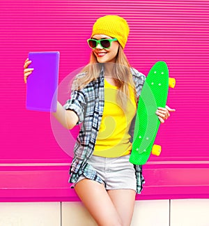 Cool smiling woman taking selfie picture by tablet pc with skateboard wearing colorful yellow hat on pink