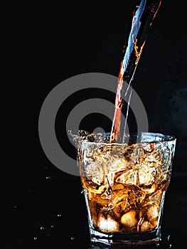 Cool, soft carbonated cola drink is poured into a glass of ice. Soda water refreshing drink isolated on dark background