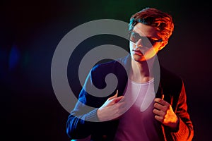 Cool stylish man in black jacket and sunglasses. High Fashion male model in colorful bright neon lights posing on black