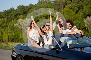 Cool stylish young dark-haired girls and guys in sunglasses smile in a black cabriolet on the road holding their hands