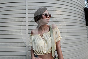 Cool stylish young hipster girl with sunglasses in a fashionable summer blouse with a bag walks on the street