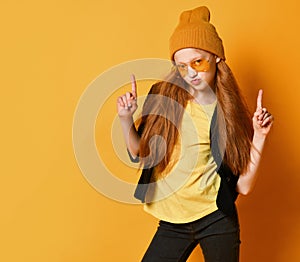 Cool teen girl with long red hair in knitted hat, yellow glasses and t-shirt is pointing fingers up like guns