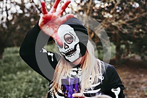 Cool teenager girl dressed up as skeleton having fun at Halloween, trick-or-treating reaching with bloody hand to camera