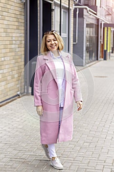 A cool young attractive woman in a pink coat and white clothes is standing on the street in the city and smiling