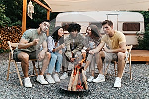 Cool young diverse friends having outdoor picnic near RV, frying marshmallows on campfire during summer vacation