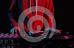 Cool young DJ in red hoodie scratching vinyl records on turntables in night club. Hands of a hip hop disc jockey playing music on