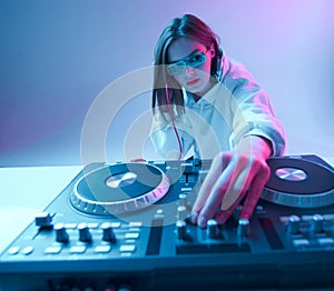 Cool young girl DJ mixes music on a mixing console and headphones, in stylish clothes, glasses on a neon background.