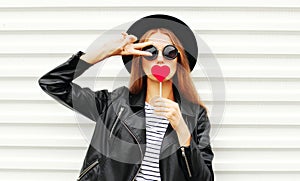 Cool young girl with red lollipop heart wearing fashion black hat leather jacket over white urban