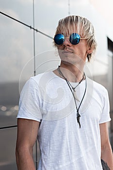 Cool young hipster man with blond hair in blue round sunglasses in a fashionable white t-shirt with amulets on a neck is standing