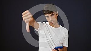Cool, young hispanic man in casual fashion delivering a negative thumb down gesture, looking unhappily at his smartphone,