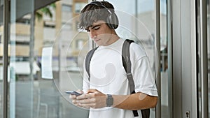 Cool young hispanic university student immersed in music, tapping on smartphone screen under campus sunlight
