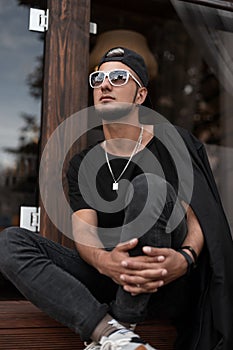 Cool young man with a beard in a fashionable black cap in a black stylish t-shirt with white fashionable sunglasses is sits