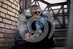 Cool young man hipster in stylish blue denim clothes in trendy sunglasses sits on a vintage staircase near brick wall in the city