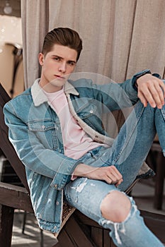 Cool young man in stylish blue denim jacket in ripped vintage jeans in pink jersey sits on street. Attractive fashionable guy in