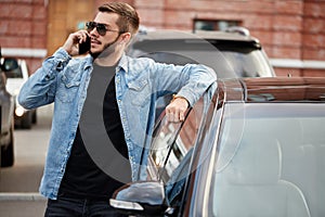 Cool young man is talking on the smart phone while standing near the car