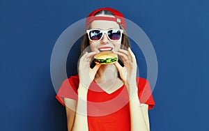 Cool young woman eating burger wearing baseball cap, sunglasses over blue wall