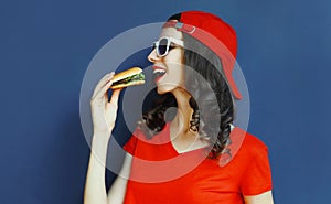 Cool young woman eating burger wearing baseball cap, sunglasses over blue wall