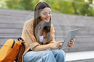 Cool young woman student sitting at park, using digital tablet