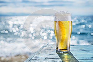 Cooled glass of beer with condensation drops on the blue wooden table. Blurred sea is at the background