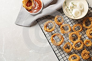 Cooling rack with homemade crunchy fried onion rings and sauces on light background, top view