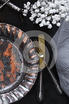 Copper/metal vintage kitchen utensils: corkscrew, vase of flowers, plate, knife, fork on black background. Still life, top view