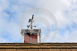 Copper rooster weathervane on top of red rooftop cupola with a blue sky and white clouds in the background