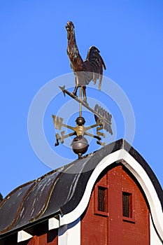 Copper weathervane on red barn cupola, blue sky background.