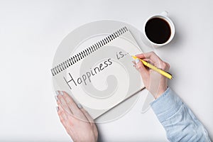 Copybook with HAPPINESS IS text. Top view of female hands writing in Notebook, cup of coffee