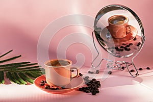 Coral colored cup with effuse coffee`s beans with shadows reflected in the mirror on pastel rose background. Coffee concept.