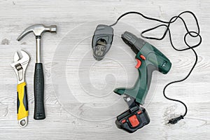 A cordless drill, it charger beside a hammer and a wrench on wooden table background.