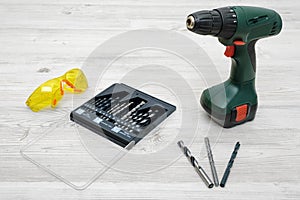 A cordless drill set to stand on a wooden table background with a set of bits in the box and yellow protective glasses