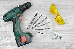 A cordless drill set on a wooden table background with a set of bits in the box and yellow protective glasses around.