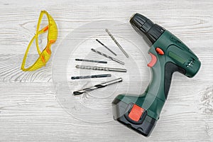 A cordless drill set on a wooden table background with a set of bits in the box and yellow protective glasses around.