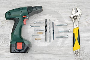 A cordless drill, a wrench, several drill bits, bolts and dowels on light wood background.
