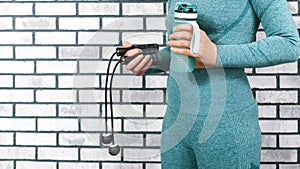 Cordless jump rope and a water bottle in the hands of a woman in sportswear on the background of a brick wall. Home fitness