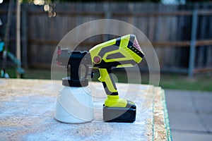 Cordless paint sprayer gun with full container of white paint on oriented strand board OSB board wood panel and blurry wooden