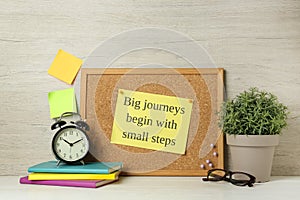 Cork board with motivational quote Big Journey Begin with Small Steps, notebooks, alarm clock and plant on white wooden table