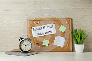Cork board with motivational quote Good Things Take Time, notebook, alarm clock and plant on white wooden table