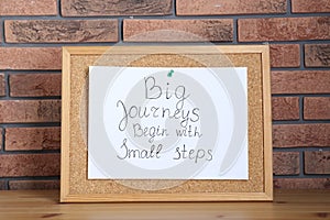 Corkboard and phrase Big Journeys Begin With Small Steps on table against brick wall. Motivational quote