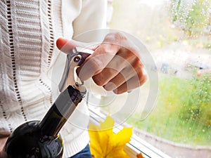 Lady sitting on the windowsill opens a bottle of wine with a corkscrew.