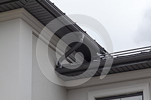 Corner of house with new gray metal tile roof and rain gutter. Metallic Guttering System, Guttering and Drainage Pipe Exterior