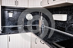 A corner with a sink in a white kitchen with a black marble countertop with a railing. Colander, sockets, knives, bottle and