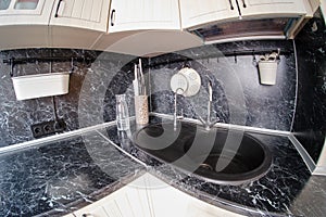 Corner with sink in a white kitchen with black marble top with railing. TV, Knives, bottle and cutlery above the table fisheye