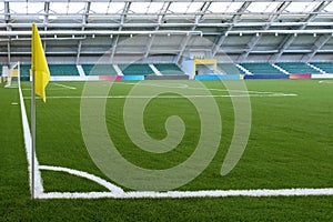 Corner of a soccer field in an indoor stadium. Yellow flag, white marking on green grass. Spectator stands in the background. Copy